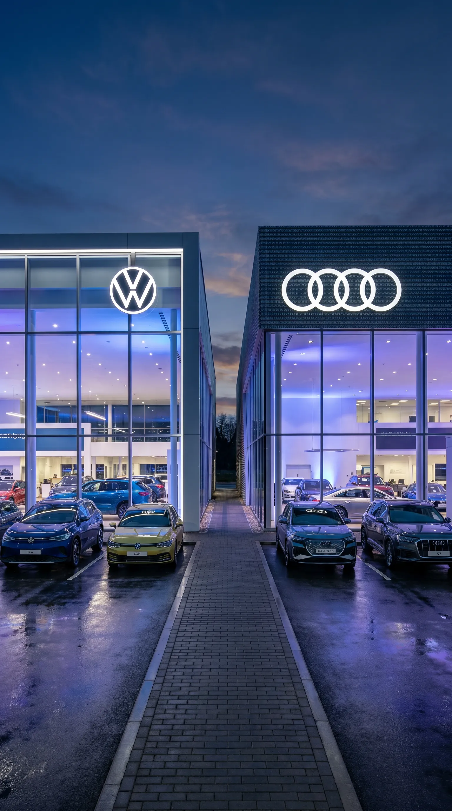 VW- und Audi-Autohaus nebeneinander bei Nacht, blau und violett beleuchtete Glas-Showrooms